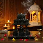 Black temple-like structure with incense and flowers on a wooden table, with a blurred background of candles and decor.